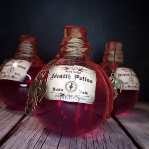 May include: Three round glass bottles filled with a vibrant pink liquid, labeled "Health Potion Bubble Bath." Each bottle has a red wax seal, twine, and a decorative charm. The bottles sit on a wooden surface against a dark background.