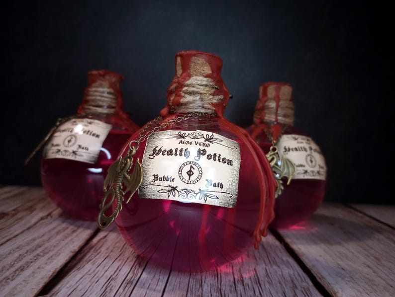 May include: Three round glass bottles filled with a vibrant pink liquid, labeled "Health Potion Bubble Bath." Each bottle has a red wax seal, twine, and a decorative charm. The bottles sit on a wooden surface against a dark background.