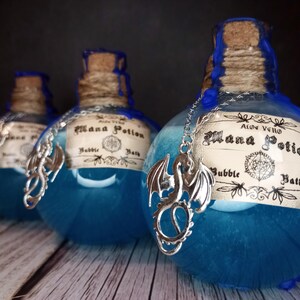 May include: Three blue glass bottles with cork stoppers and silver dragon charms. Each bottle has a label that reads "Mana Potion Bubble Bath". The bottles are filled with a blue liquid and are decorated with blue wax.