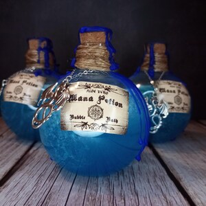 May include: Three blue potion bottles with cork stoppers and decorative labels. The bottles are adorned with silver charms and twine. The labels read "Mana Potion Bubble Bath" and "Aloe Vera". The bottles are arranged on a wooden surface.