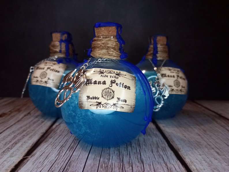 May include: Three blue potion bottles with cork stoppers and decorative labels. The bottles are adorned with silver charms and twine. The labels read "Mana Potion Bubble Bath" and "Aloe Vera". The bottles are arranged on a wooden surface.