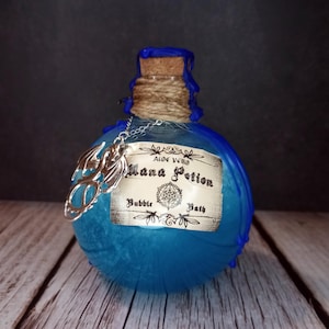 May include: A blue glass bottle with a cork stopper, twine, and a silver charm. The label reads "Mana Potion Bubble Bath." The bottle has a textured surface and is set against a dark background on a wooden surface.