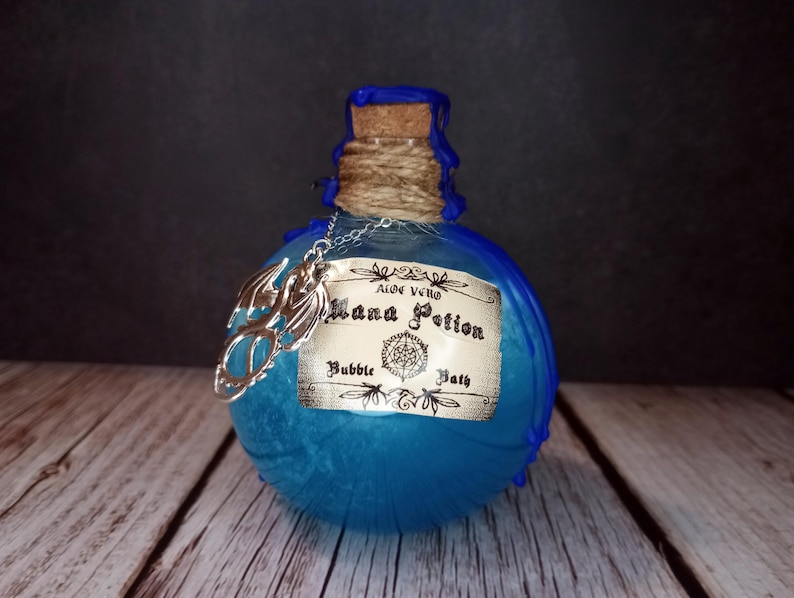 May include: A blue glass bottle with a cork stopper, twine, and a silver charm. The label reads "Mana Potion Bubble Bath." The bottle has a textured surface and is set against a dark background on a wooden surface.