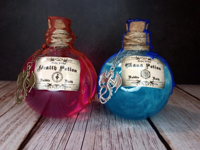 May include: Two potion bottles with cork stoppers and dragon charms. One contains pink liquid, labelled "Health Potion Bubble Bath." The other has blue liquid, labelled "Mana Potion Bubble Bath." The bottles are on a wooden surface.