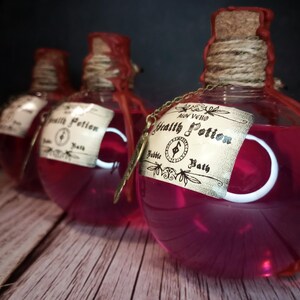 May include: Three round glass bottles filled with a vibrant pink liquid, labeled "Health Potion Bubble Bath." Each bottle has a cork stopper, twine, and a decorative tag. The bottles are on a wooden surface against a dark background.