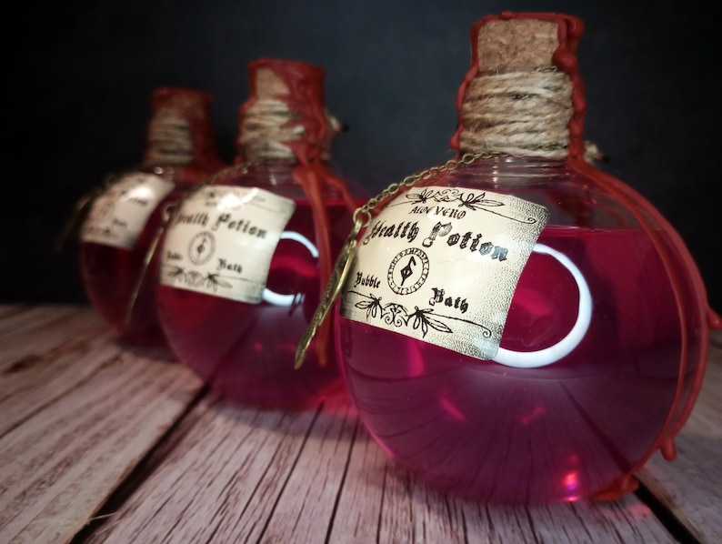May include: Three round glass bottles filled with a vibrant pink liquid, labeled "Health Potion Bubble Bath." Each bottle has a cork stopper, twine, and a decorative tag. The bottles are on a wooden surface against a dark background.