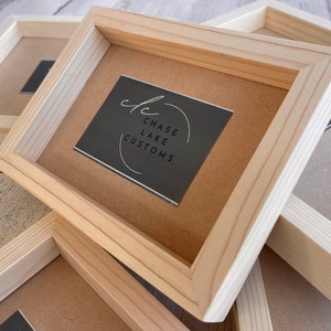 May include: Close-up of several unfinished wooden picture frames stacked on top of each other. The top frame has a black card inside with the text "Chase Lake Customs" in white lettering.