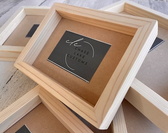 Puede incluir: Primer plano de varios marcos de fotos de madera sin terminar apilados uno encima del otro. El marco superior tiene una tarjeta negra en el interior con el texto "Chase Lake Customs" en letras blancas.
