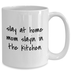 May include: White ceramic coffee mug with black text that reads "slay at home mom slayin in the kitchen".