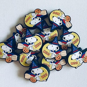 May include: A collection of Halloween-themed Snoopy shoe charms. Each charm features Snoopy dressed as a witch, holding a broom and a pumpkin, with the word "BOO" on a yellow background. The charms have a black border and are scattered on a white surface.