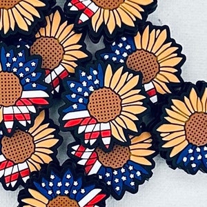 May include: A collection of sunflower-shaped charms with a patriotic design. Each charm features a yellow sunflower with a brown center and a section of the American flag, including red and white stripes and blue stars.