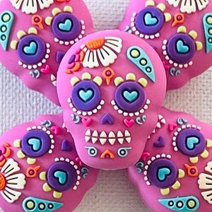 May include: Five pink sugar skull decorations arranged in a flower shape. Each skull features intricate details, including heart-shaped eyes, floral accents, and a row of white teeth. The skulls are adorned with purple, blue, orange, and white details.
