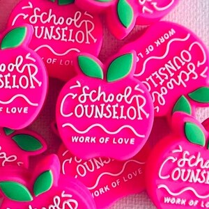 May include: A collection of pink apple-shaped charms with green leaves. Each charm has the words "School Counselor" and "Work of Love" in white lettering. The charms are scattered on a white surface.