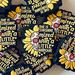 May include: A collection of black shoe charms with a sunflower and skull design. The charms are yellow and black, with the text "She is sunshine mixed with a little hurricane". The charms are scattered on a white surface.