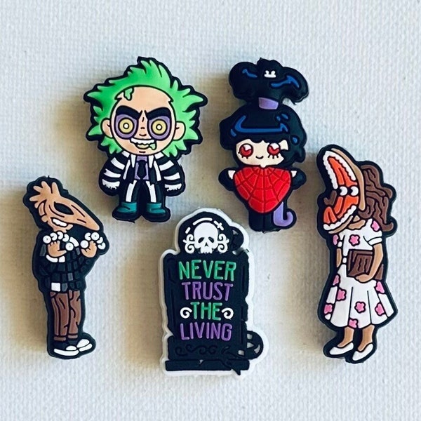 May include: Set of five colorful cartoon character shoe charms featuring a green-haired character with a black and white striped shirt, a red-haired character with a white dress and a red heart, a brown-haired character with a white dress and a book, a brown character with a black and white shirt and a white collar, and a tombstone with the text "Never Trust The Living".