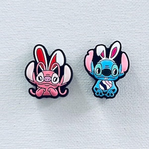 May include: Two blue and pink cartoon character shoe charms with bunny ears. The blue character has a white and black striped egg. The pink character has red and white bunny ears.