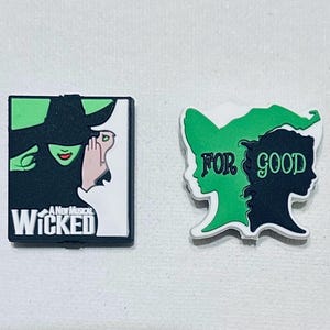 May include: Two Wicked musical themed shoe charms. One charm features a green witch with a black hat and the text "A New Musical Wicked". The other charm features a green and black silhouette of two women with the text "For Good".
