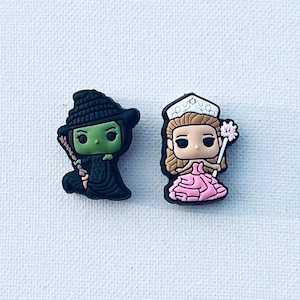 May include: Two shoe charms featuring characters from The Wizard of Oz. One charm is a green-skinned witch in a black hat and dress, holding a staff. The other is a princess in a pink dress, holding a flower and wearing a crown.