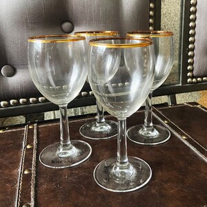 May include: Four clear glass wine glasses with gold rims sit on a brown leather surface. The glasses are empty and appear to be new.