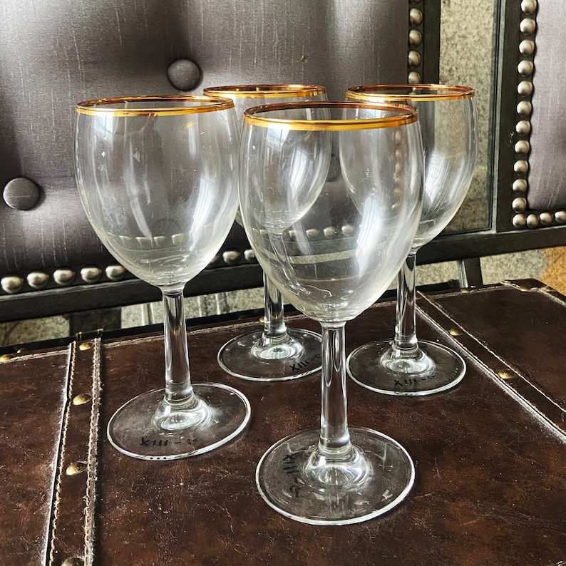 May include: Four clear glass wine glasses with gold rims sit on a brown leather surface. The glasses are empty and appear to be new.