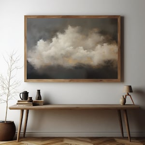 Puede incluir: Una pintura enmarcada de una gran formación de nubes blancas sobre un cielo gris oscuro. La pintura está colgada en una pared blanca sobre una mesa de madera con un jarrón, una jarra y una lámpara.