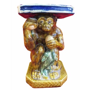 May include: A ceramic monkey stool with a blue and white glazed top. The monkey is brown and has a green leaf in its hand. The stool is sitting on a woven base.