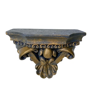 May include: Ornate wall shelf with a dark gray top and gold-colored decorative accents. The shelf features intricate scrollwork and a stylized floral design, creating a vintage aesthetic. The shelf is designed to be mounted on a wall.
