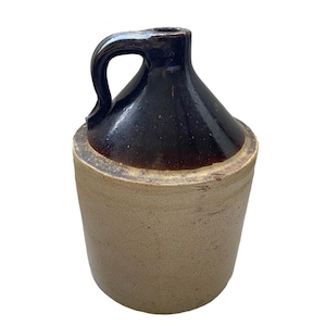 May include: A brown and tan stoneware jug with a handle. The jug has a dark brown glaze on the top half and a light tan glaze on the bottom half.
