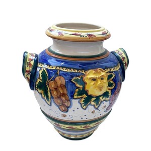 May include: A white ceramic vase with blue, green, yellow, and red painted details. The vase has two handles and features a grapevine design with a stylized face.