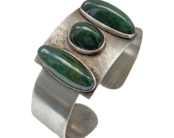 May include: A silver cuff bracelet featuring three oval-shaped, dark green gemstones. The gemstones are set in a textured silver plate, with the bracelet's band having a smooth, polished finish. The design is bold and modern.