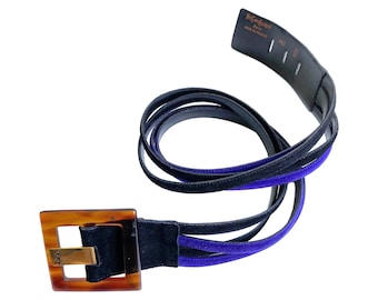 May include: A black and blue belt with a square tortoiseshell buckle. The belt has a gold-colored metal accent on the buckle and the brand name "Yves Saint Laurent" is visible on the belt strap.
