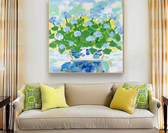May include: A framed painting depicts a vibrant still life of blue hydrangeas in a white and blue patterned vase. The artwork features a gold frame and is displayed above a beige sofa with yellow and green throw pillows.