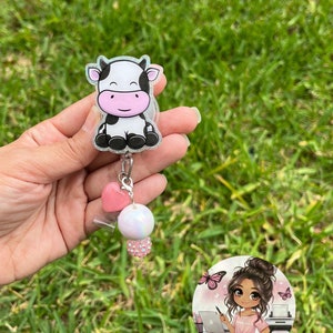 May include: A black and white cow cartoon charm on a clear retractable badge holder with a pink heart and white pearl charm. The charm is attached to a silver chain. The background is green grass.