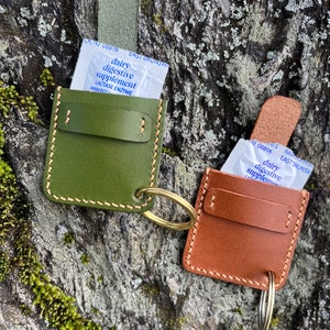 May include: Two leather keychains with a small pocket for holding packets of dairy digestive supplement. The keychain on the left is green and the keychain on the right is brown.