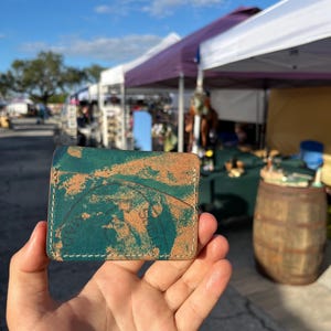 May include: A teal leather wallet with a distressed, aged look. The wallet has a brown leather interior and a white stitch around the edges.