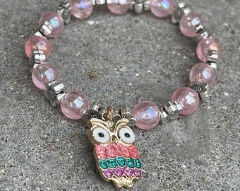 May include: Pink beaded bracelet with a silver owl charm. The owl has sparkly pink, green, and white details.