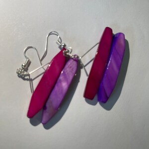 Könnte beinhalten: Ein Paar Ohrringe mit einem mehrschichtigen Design. Die Ohrringe sind aus lila und rosa Achat und haben silberne Haken.