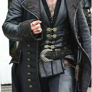 Puede incluir: Un abrigo largo de cuero negro con botones dorados y un chaleco a juego. El chaleco tiene cierres dorados decorativos. El atuendo incluye un cinturón ancho con hebilla plateada y una funda. La persona lleva pantalones grises y un collar.