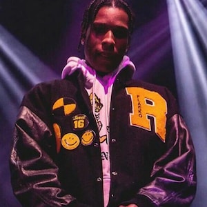 May include: A black and yellow varsity jacket with a large yellow "R" on the back. The jacket has a white hoodie underneath and features various patches, including a smiley face and a "Sweet 16" patch.