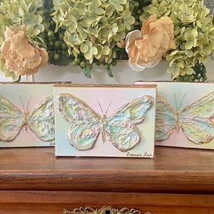 May include: Five small canvases with painted butterflies in shades of blue, green, and gold. Each canvas has a different background color, including pink, blue, and green. The canvases are arranged in a row on a wooden surface.