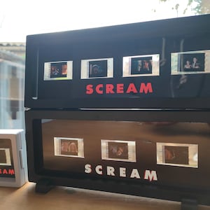 May include: Black framed display featuring film strips from the movie "Scream." The word "SCREAM" is displayed in red block letters. A smaller white framed display with the word "SCREAM" in red is also visible.