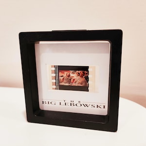 Könnte beinhalten: Ein schwarz gerahmtes Display mit einem Filmstreifen mit einer Szene aus "The Big Lebowski". Der Filmstreifen zeigt drei Personen in einem Raum. Der Text "THE BIG LEBOWSKI" ist unten im Rahmen weiß gedruckt.