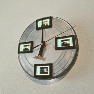 May include: A unique wall clock made from a silver film reel. The clock features four small framed images and black clock hands. The clock hands are black with a gold center. The clock is mounted on a white wall.