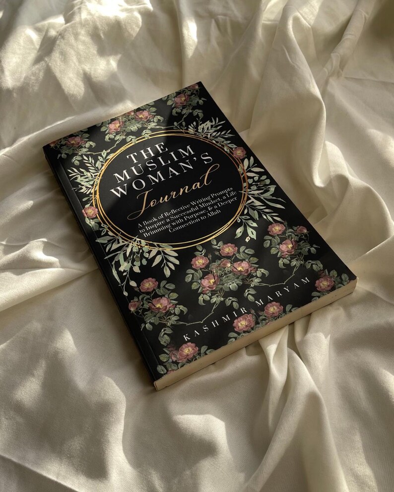 May include: A black journal with gold lettering that reads "The Muslim Woman's Journal". The cover features a floral design with pink roses and green leaves.