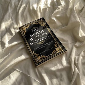 May include: A black book with gold lettering titled "The Muslim Woman's Manifesto: 10 Steps to Achieving Phenomenal Success, in Both Worlds" by Kashmir Maryam, MIT.