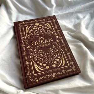 May include: A brown journal with gold floral designs and the text "My Quran Journal." The cover features an ornate border and the name "Kashmir Marvam" at the bottom. The journal is set against a white, textured background.