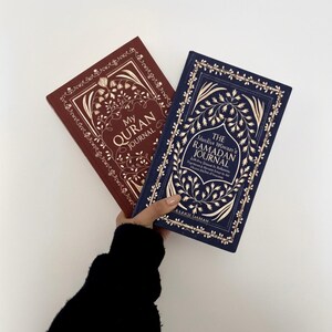 May include: Two hardcover journals, one burgundy and one navy blue, with gold floral designs and text. The burgundy journal reads "My Quran Journal," and the navy journal reads "The Ramadan Journal."
