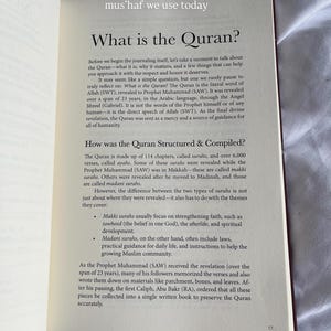 May include: Open book with text about the Quran, including its revelation and compilation. The pages are white with black text, and the book is open to a section titled "What is the Quran?" and "How was the Quran Structured & Compiled?"