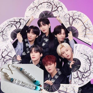 May include: A pair of silver earrings with white beads spelling out "ONEUS" and "TOMOON" with blue and red beads. The earrings are shown in front of a group of six men wearing black and white traditional clothing and holding white fans.