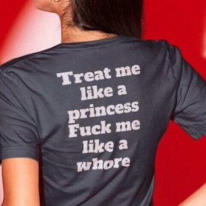 May include: A dark gray t-shirt with the text "Treat me like a princess, Fuck me like a whore" in white. The person is wearing black leather pants. The background is red.